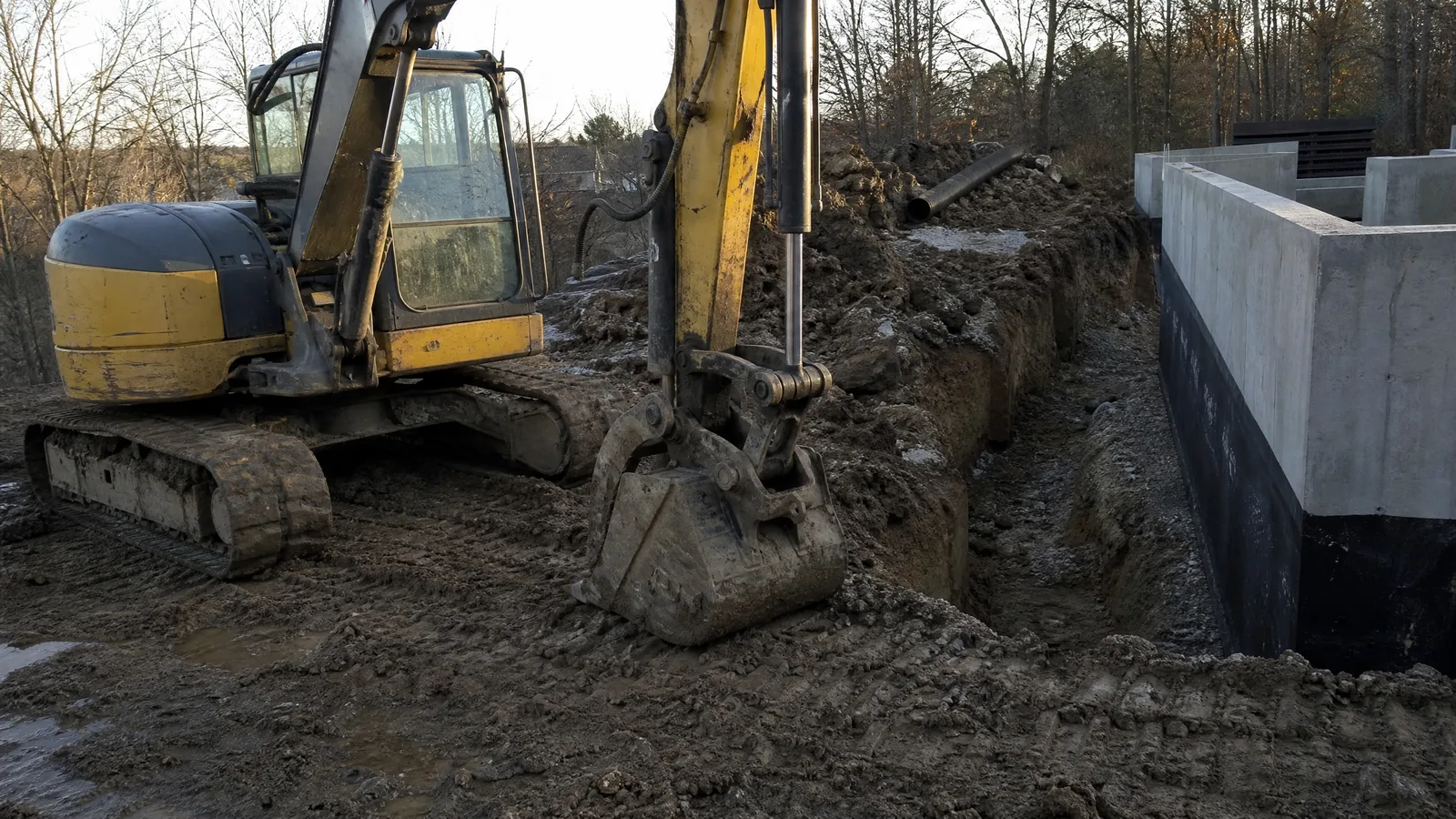 Excavator bucket resting at the edge of an open trench beside a poured concrete foundation wall